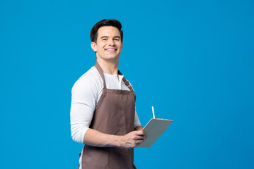 Fototapeta na wymiar Caucasian man with apron holding writing pad and pen in his hands while standing on blue background in light studio