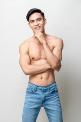 Fototapeta premium Smiling handsome muscular Asian man with black hair and naked torso touching chin while standing on gray isolated background in light studio