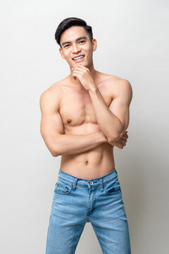 Smiling handsome muscular Asian man with black hair and naked torso touching chin while standing on gray isolated background in light studio