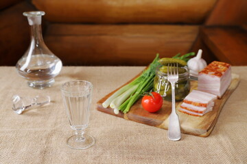 A glass of vodka is on the table next to pickles, fresh greens, vegetables, bacon and a decanter on...