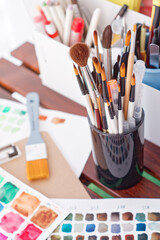 Various brushes are in a holder next to a sheet of paper with hand-painted watercolor palettes in the form of square spots. Creative concept. Preparation of paints for creativity.