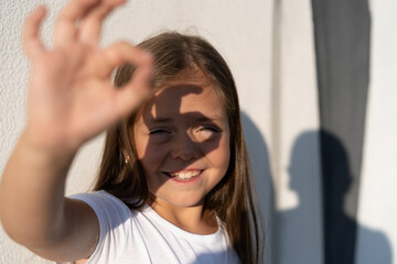 Fashion kid concept - portrait of a girl covering her face from the bright sun while standing against the white wall.