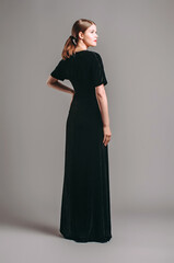 Black velvet trumpet dress. Evening floor length gown with deep v neck line and short sleeves. Beautiful young brunette lady with red lips and ponytail hairstyle posing in studio. Total black look.