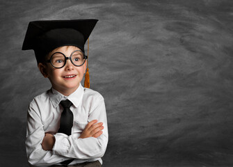 Happy Child in Glasses and Graduation Hat thinking over Blackboard. Cheerful smiling Preschool Boy...
