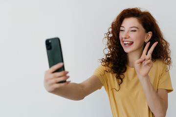 Beautiful redhead woman with curly hair, touching her hairstyle, smiling and taking selfie, making photo on smartphone on white background