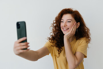 Beautiful redhead woman with curly hair, touching her hairstyle, smiling and taking selfie, making photo on smartphone on white background