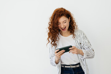 Young redhead woman playing video games on her phone, watching video live stream on cellphone, laughing and smiling, looking at mobile screen, white background