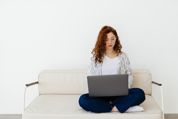 Beautiful red-haired woman working online, using laptop, sitting on comfortable grey couch