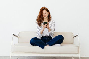 Ginger young woman lying on sofa, using online app on modern smartphone, spending weekend day at home