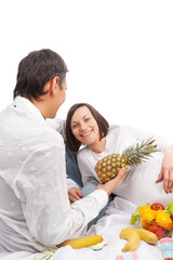 Pregnancy Concepts. Funny Portrait of Caucasian Husband Man Offering His Pregnant Wife Fresh Pineapple Over White Background.