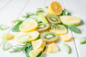 Partially blurred green and yellow fruits and herbs cut into pieces lie on white table. Ingredients for smoothie, fruit salad or dessert. Slices of lemon, apple, kiwi, cucumber, spinach and tarragon