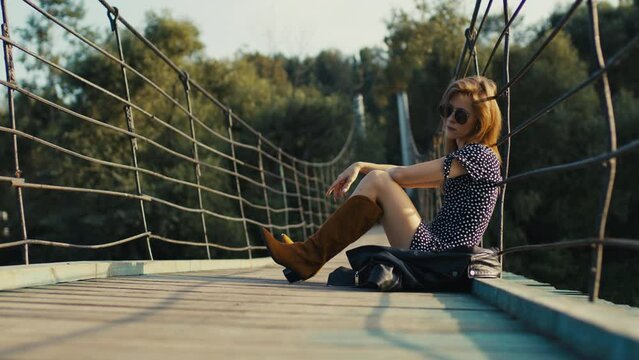 A young, girl in a dress and cowboy boots sits on a suspension bridge at sunset.
