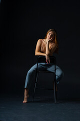 Naklejka premium Sexy blonde girl model in a pink bra on a bare torso and gray trousers posing barefoot sitting on a chair in the studio on a black background in provocative poses. Seduction in the dark