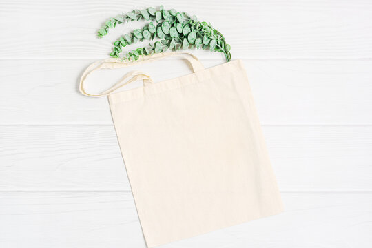 Beige blank template as commercial shopping tote bag canvas and green eucalyptus plant branch on white wood background. Flatlay, copyspace, top view.
