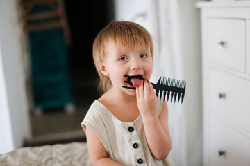 Funny baby toddler in baby overalls nibbles on a comb in bedroom, child safety and teething, dirt...