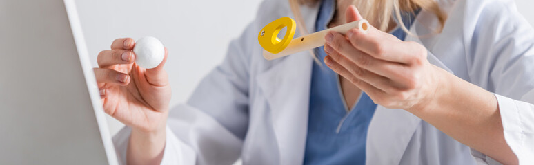 Cropped view of speech therapist in white coat holding respiratory muscle trainer near laptop in consulting room, banner.