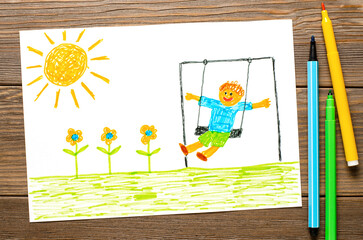 A boy swings on a swing at noon. Children's drawing with felt-tip pens. Wooden table