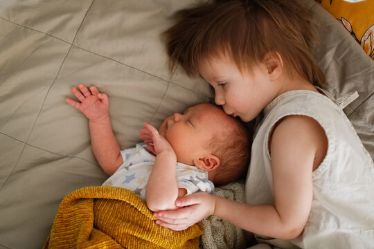 cute chubby baby sleeps on bed next to an older sibling, children are siblings together. older sister kisses and hugs the baby. In real cozy interior