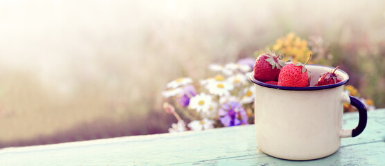 red strawberries in a metal light mug on an old table in a field against the background of a sunset. summer retro dessert