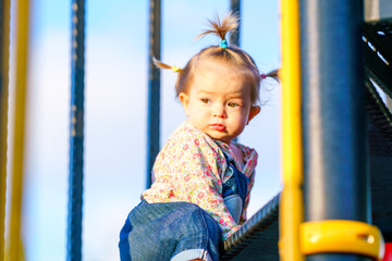 The kid is learning to climb stairs. Baby has multi-colored short pigtails. Happy child is playing on the playground. Active games for children. Funny girl with pigtails on a children's slide