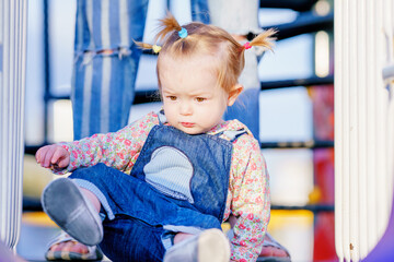 Fototapeta premium Happy child is playing on the playground. Funny girl with pigtails on a children's slide. Baby has multi-colored short pigtails. The kid is learning to climb stairs. Active games for children