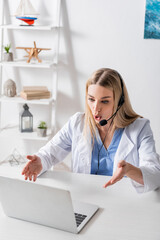 Speech therapist in headset having video call on laptop in consulting room.