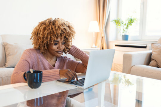 A young woman enjoys a modern room at home. A beautiful young Afro woman sitting in the living room, uses a laptop, drinks coffee and enjoys her free time at home.