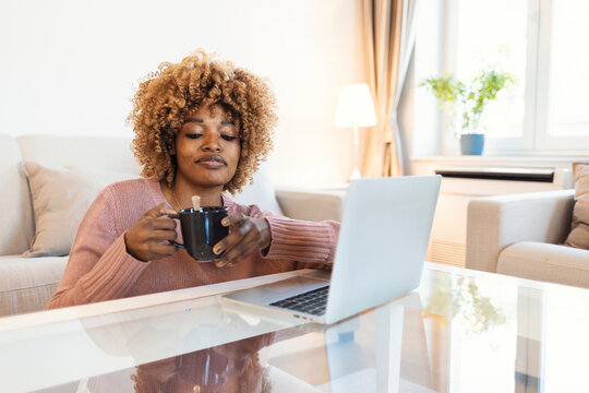 A young woman enjoys a modern room at home. A beautiful young Afro woman sitting in the living room, uses a laptop, drinks coffee and enjoys her free time at home.