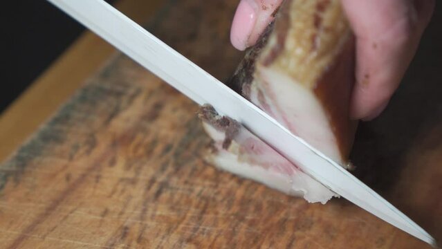 A woman's hand, with a knife, cuts a slice of lard on a wooden cutting board.
