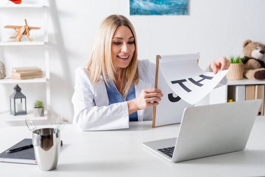Cheerful logopedist holding clipboard with letters during video call on laptop in consulting room.