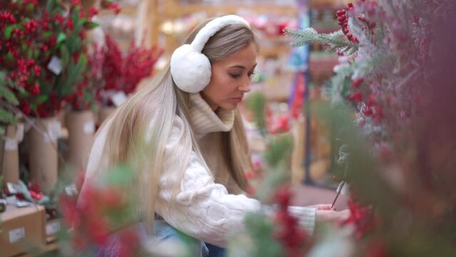 Christmas shopping, European woman choose branches for Christmas wreath indoor in supermarket. Caucasian female buying Christmas decor on holiday market, dressed fur headphones and white sweater. Sale