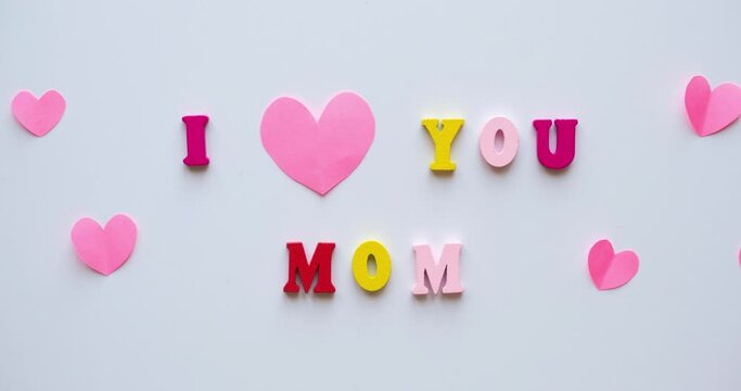 Phrase I Love You Mom from colorful wooden letters and a paper pink hearts flying from bottom to top. Congratulations on mother's day. 4K looped stop motion animation