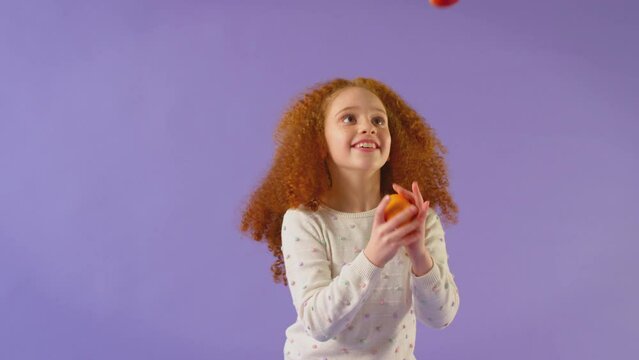 Studio Portrait Of Girl Juggling Apple And Orange Against Purple Background