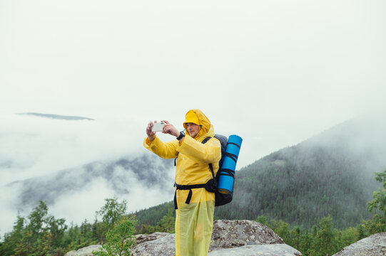 Hiker in a raincoat with a backpack on his back stands on top of a mountain against the backdrop of a landscape on a forest in the fog and takes photos on a smartphone camera.