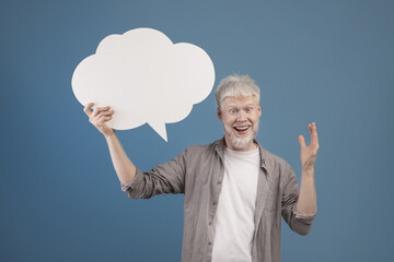 Happy albino man holding blank speech bubble and smiling at camera, posing over blue background,...