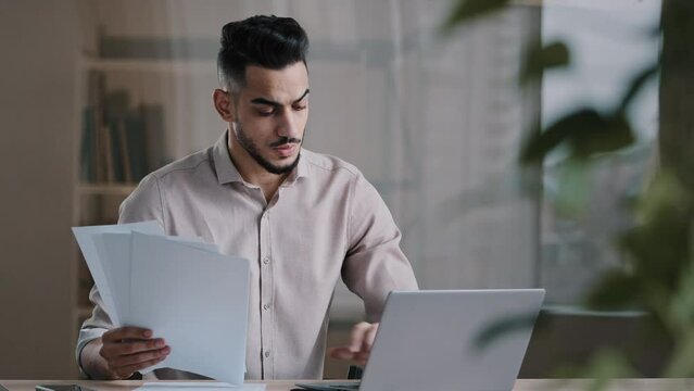 Serious arabian young business guy male professional worker employee type on computer keyboard copy paper text at electronic form focused hispanic man compare documents check online financial report
