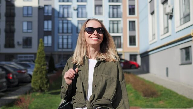 Portrait of young attractive smiling woman in sunglasses looking at camera on a background of colored city buildings. Portrait happy female tourist having good time wandering around town in summer.