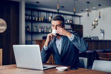 A businessman is sipping his coffee while looking at the laptop. 