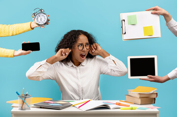Young black woman sitting at desk surrounded by hands with gadgets and work related objects, covering ears and screaming