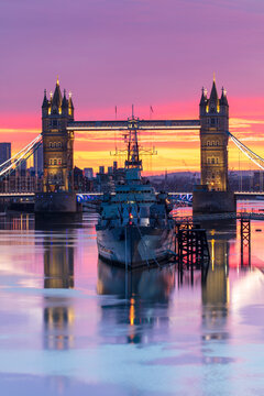Tower Bridge and HMS Belfast reflecting in a still River Thames at sunset, London