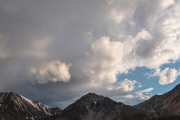 Obraz premium Dramatic mountain landscape with high mountain range with sharp rocky pinnacle under clouds of sunset color in gloomy sky. Dark atmospheric scenery with large mountains with snow at changeable weather
