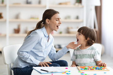 Professional woman speech therapist helping little boy to pronounce right sounds, showing mouth...