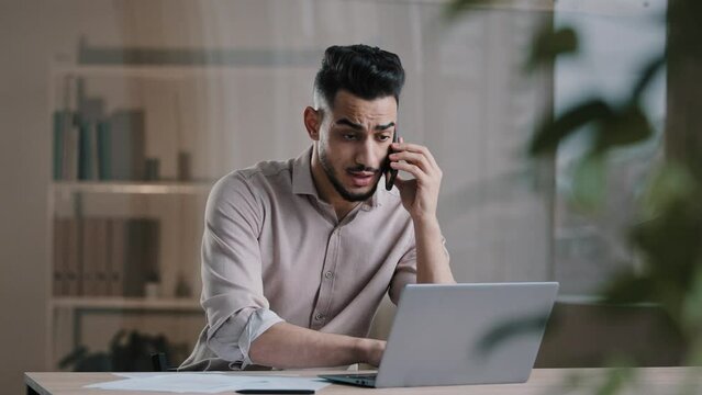 Busy millennial arabian business man web designer employee using laptop talking on cell phone involved in multitasking using gadgets discussing online project distant noting paper documents at office