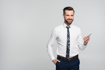 cheerful businessman holding hand in pocket and mobile phone isolated on grey.