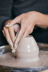 Female hands sculpt clay dishes. Сraftswoman in apron sitting at pottery wheel and using craft tool while shaping wet clay vessel