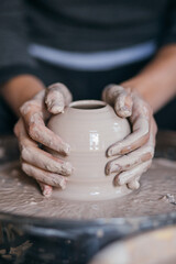 Female hands sculpt clay dishes. Сraftswoman in apron sitting at pottery wheel and using craft tool while shaping wet clay vessel