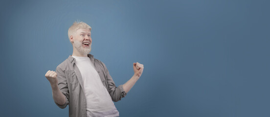 Great news. Overjoyed albino guy screaming and shaking clenched fists, posing over blue background,...
