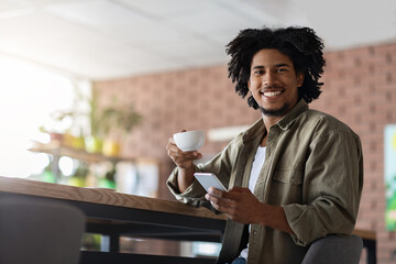 Cheerful millennial african american curly man with smartphone sit at table, drink cup of coffee
