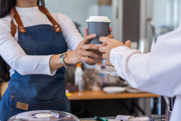 young barista delivers hot coffee in take away paper cup to customer before making payment at...