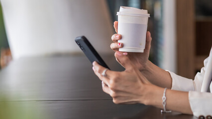 businesswoman with paper cup of hot coffee and mobile phone connected to Internet to trade stocks through an app during break in coffee shop.Drinking coffee concept to help stimulate brain.